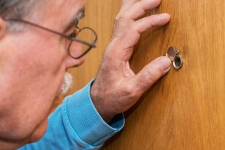 Starší muž vyhlíží kukátkem exekutora za dveřmi