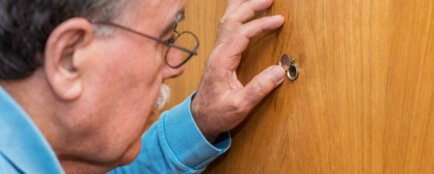 Starší muž vyhlíží kukátkem exekutora za dveřmi