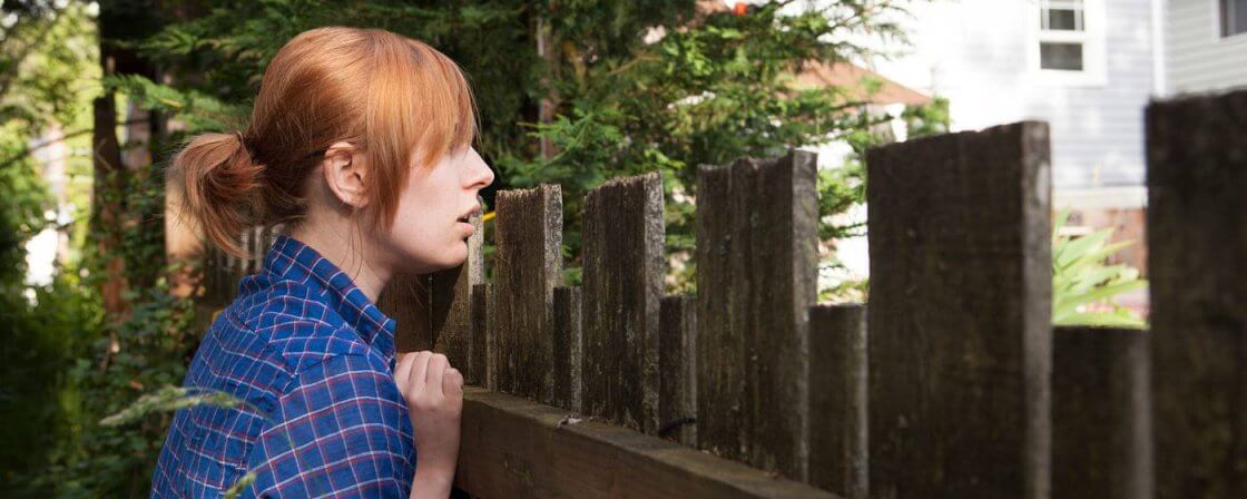 Žena stojící u vysokého plotu se dívá na sousedovu zahradu